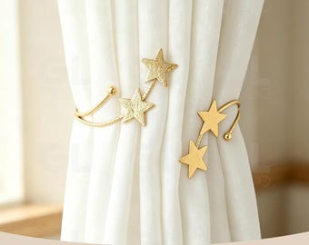 May include: Two gold-toned metal curtain tiebacks featuring star-shaped accents. One tieback displays three textured stars, while the other features two smooth stars. Both pieces are curved to hold white fabric curtains in place.