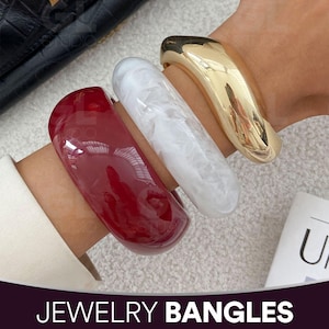 May include: Three bold bangles in red, white, and gold. The red bangle is a glossy, solid colour. The white bangle has a marbled effect. The gold bangle is smooth and reflective. The text "JEWELRY BANGLES" is at the bottom.
