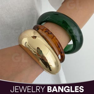May include: Three bangles in gold, tortoiseshell, and green are stacked on a wrist. The gold bangle is a wide, smooth, and reflective band. The tortoiseshell bangle has a marbled pattern. The green bangle is a solid, translucent color. The text "JEWELRY BANGLES" is at the bottom.