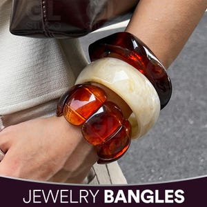 以下が含まれることがあります： 琥珀色、クリーム色、濃い茶色の太いバングルのコレクション。ジュエリーには、幅広の大理石模様のクリーム色のバングル、濃い茶色の角張ったバングル、琥珀色の丸みを帯びたバングルのクラスターが含まれています。「JEWELRY BANGLES」というテキストが下にあります。