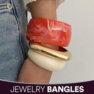Puede incluir: Tres pulseras apiladas. La pulsera superior es de forma rectangular ancha, roja con vetas blancas. Debajo, una pulsera dorada de forma cuadrada redondeada y en la parte inferior, una pulsera crema de forma cuadrada redondeada. El texto "JEWELRY BANGLES" está en la parte inferior.