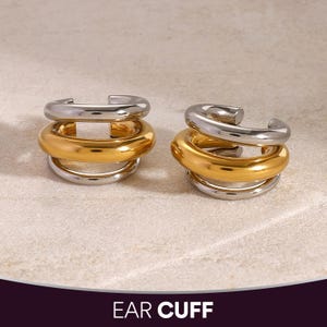 Könnte beinhalten: Zwei Ohrklemmen, jeweils mit einem silbernen oberen und unteren Band und einem goldfarbenen mittleren Band. Die Ohrklemmen sind gestapelt und haben ein modernes, minimalistisches Design. Der Text "EAR CUFF" befindet sich am unteren Bildrand.