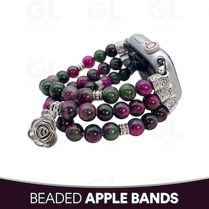 Peut inclure: Un bracelet de montre composé de plusieurs rangs de perles polies dans des tons magenta profond et vert forêt. Le bracelet comprend des entretoises en métal argenté et une breloque rose argentée, fixée à un connecteur de montre intelligente.