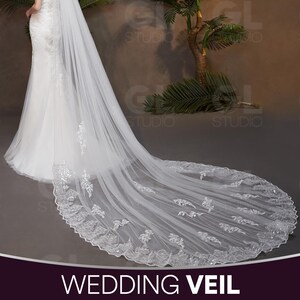 May include: A long, flowing white wedding veil with intricate lace detailing along the edges. The veil is draped over a white wedding dress, creating a graceful and elegant look. The text "WEDDING VEIL" is displayed at the bottom of the image.