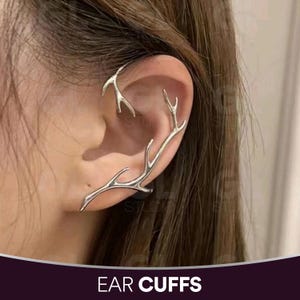 May include: Silver-toned ear cuffs in an antler design. The ear cuffs curve around the ear, with the text "EAR CUFFS" in white at the bottom of the image. The jewelry is worn on a person's ear.