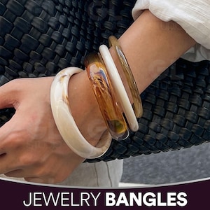 Może przedstawiać: Na zdjęciu widoczne są cztery bransoletki w różnych kolorach i wzorach. Bransoletki obejmują kremowy marmurkowy wzór, przezroczystą bursztynową bransoletkę, białą bransoletkę i jasno brązową bransoletkę. Tekst "JEWELRY BANGLES" znajduje się na dole obrazu.