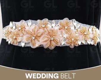 May include: A bridal belt featuring a grey fabric base, embellished with peach fabric flowers, gold beads, and clear crystal accents. The belt is decorated with pearls and clear, teardrop-shaped crystals. The text "WEDDING BELT" is at the bottom.
