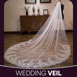 May include: A long, white wedding veil with an ornate, embroidered border. The veil is displayed on a mannequin and drapes across the floor. The bottom edge features detailed floral and beaded embellishments. The text "WEDDING VEIL" is at the bottom of the image.