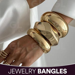 Könnte beinhalten: Goldfarbene Armreifen und Ringe. Das Bild zeigt eine Hand, die mehrere klobige, polierte goldene Armreifen und Ringe trägt. Der Schmuck wird vor einem weißen Hintergrund präsentiert. Der Text "JEWELRY BANGLES" befindet sich am unteren Bildrand.