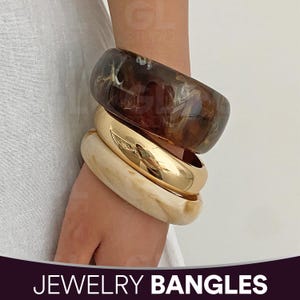Puede incluir: Tres pulseras apiladas. La pulsera superior es de color marrón y crema veteado. La pulsera del medio es dorada. La pulsera inferior es de color crema claro. El texto "JEWELRY BANGLES" está en la parte inferior de la imagen.