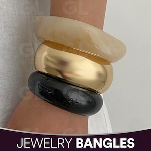 Può includere: Tre braccialetti impilati in crema, oro e nero. Il braccialetto color crema è traslucido, quello dorato è lucido e quello nero è lucido. L'immagine include il testo "JEWELRY BANGLES" in basso.
