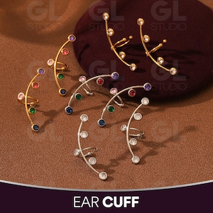 May include: Ear cuffs in gold and silver tones, adorned with colorful and clear gemstones. The ear cuffs curve along the ear, with a ring that fits behind the ear. The text "EAR CUFF" is at the bottom of the image.