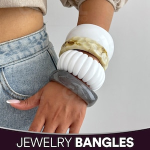 May include: A set of four stacked bangles worn on the wrist. The collection includes a smooth white bangle, a marbled beige and brown bangle, a white ribbed bangle with vertical ridges, and a smooth dark gray marbled bangle.