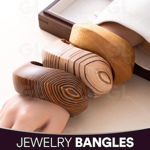 May include: Three wooden bangles in shades of brown. The bangles feature unique grain patterns and geometric designs. One bangle is worn on a wrist. A jewelry box is also visible. The text "JEWELRY BANGLES" is at the bottom of the image.
