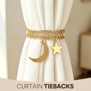 May include: A gold-toned metal curtain tieback featuring a series of interconnected circular rings forming a band. Two flat, gold-colored charms hang from the band: one in the shape of a crescent moon and the other in the shape of a five-pointed star.