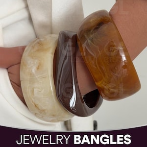 May include: Three chunky bangles in cream, dark brown, and amber hues. The cream bangle has a marbled effect, the dark brown is solid, and the amber has a swirling pattern. The text "JEWELRY BANGLES" is at the bottom.