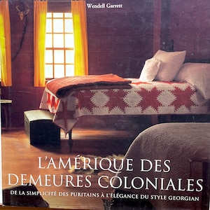 Peut inclure: Couverture de livre avec le titre "L'AMÉRIQUE DES DEMEURES COLONIALES" et l'auteur Wendell Garrett. L'image montre une chambre avec un lit recouvert d'une courtepointe à motifs d'étoiles rouges et blanches, une couverture rouge et des oreillers blancs. La pièce a des murs en bois et une fenêtre avec des rideaux orange.