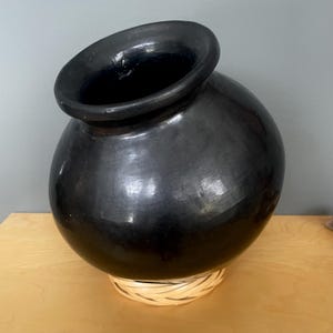 May include: A large, black ceramic pot with a wide, angled opening. The pot has a rounded body and sits on a woven, light-coloured base. The glossy surface reflects light, highlighting its smooth texture. This decorative piece is suitable for home decor.