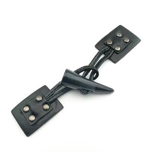 May include: Black toggle clasp with a horn-shaped closure. The clasp features two rectangular leather pieces, each with four silver-colored rivets. The horn is connected to the leather pieces by black leather cords.
