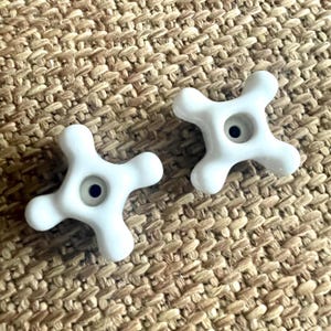 May include: Two white, cross-shaped ceramic knobs with a small, dark blue center. The knobs have a glossy finish and are set against a textured, tan background. These decorative knobs are likely for furniture or cabinetry.