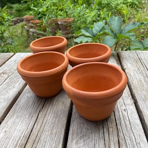 Puede incluir: Un conjunto de cuatro macetas de terracota con borde redondeado y acabado liso sin esmaltar. Cada maceta presenta una abertura ancha y una base cónica, mostrando el color marrón rojizo natural de la arcilla típica de la cerámica.