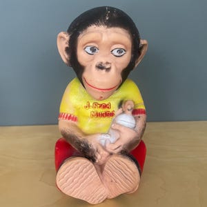 Może przedstawiać: Figurka małpy w stylu vintage, siedząca z butelką dla dziecka. Małpa ma na sobie żółtą koszulę z czerwonymi wykończeniami i napisem "J. Fred Mudd's". Małpa ma czarne futro na głowie i czerwone spodnie. Figurka jest wykonana z twardego plastiku.