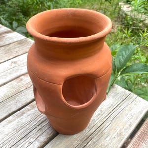 May include: A terracotta strawberry planter pot with a wide circular opening at the top and multiple smaller circular openings along the sides. The vessel is made of unglazed reddish-brown clay with a smooth, matte finish and a flared rim.