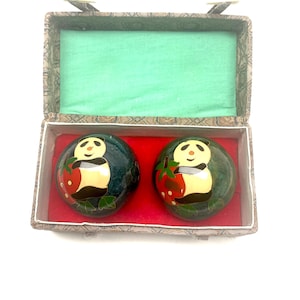 May include: Two round, dark green decorative stones featuring panda bear and red apple designs, displayed in an open rectangular box. The box has a green interior and a patterned exterior, showcasing the contents.