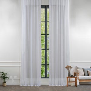 Puede incluir: Cortinas blancas y transparentes colgadas en una ventana con vista a árboles verdes afuera. Las cortinas están hechas de una tela ligera y tienen una textura sutil.