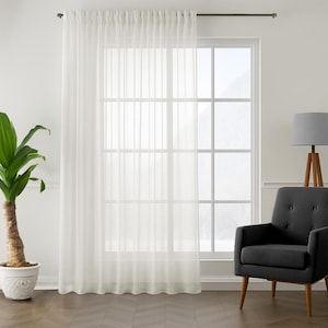 May include: Sheer white curtain panel hanging in front of a large window. The curtain is semi-transparent, allowing light to filter through. A dark gray armchair and a floor lamp with a gray shade are to the right. A potted plant is on the left.