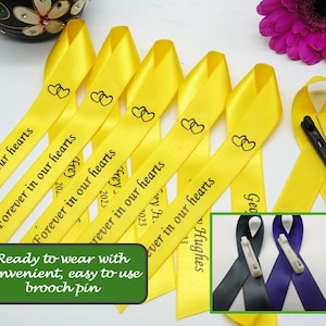 May include: Yellow satin ribbon with the text "Forever in our hearts" printed on it. The ribbon is tied in a bow and has a safety pin attached. The ribbon is ready to wear with a convenient, easy-to-use brooch pin.