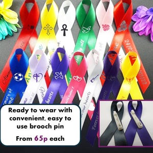 May include: A collection of colourful satin awareness ribbons featuring various printed symbols and text, including crosses, hearts, doves, and sports icons. Each ribbon includes a brooch pin on the back for attachment. The ribbons are displayed alongside artificial flowers.