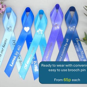 May include: Five satin ribbon pins in shades of blue, each featuring a loop at the top and a long tail. The ribbons are printed with various symbols including a dove, heart, star, and butterfly, along with memorial text such as Always in our hearts.