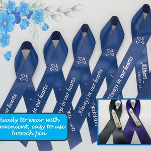 May include: Blue satin ribbon with the text "Always in our hearts" printed in white. The ribbon is tied in a bow and has a small brooch pin attached. The ribbon is a memorial ribbon.