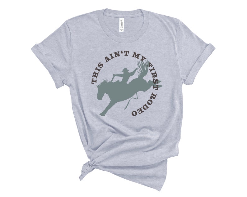 May include: A light gray t-shirt with a brown circle graphic that says "This ain't my first rodeo" with a silhouette of a cowboy riding a bucking bronco inside the circle.