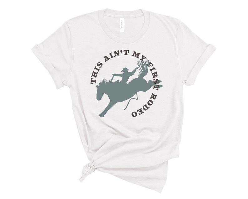 May include: White t-shirt with a gray silhouette of a person riding a bucking horse. The text "This ain't my first rodeo" is written in a circle around the image.
