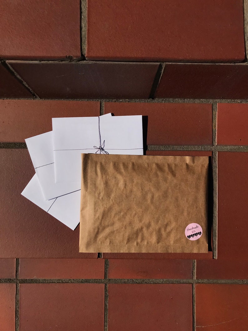 May include: A brown paper bag with a pink circle sticker that says "Handmade with love" in black text. The bag is tied with a string and has three white envelopes inside.