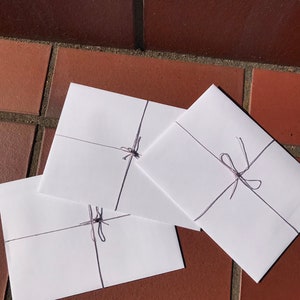 May include: Three white envelopes tied with a thin, light purple string. The envelopes are stacked on top of each other, with the top envelope slightly overlapping the middle envelope.