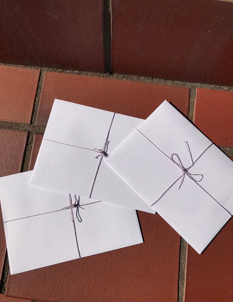 May include: Three white envelopes tied with a thin, light purple string. The envelopes are stacked on top of each other, with the top envelope slightly overlapping the middle envelope.