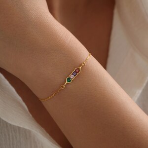 Op de afbeelding: Een gouden kettingarmband met een erwtendoppenbedel met vijf gekleurde edelstenen. De bedel bevindt zich in het midden van de armband.