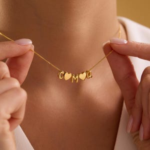 以下が含まれることがあります： C、O、M、Qの文字が入ったゴールドカラーのネックレス。Oの文字はハート型です。ネックレスは繊細なチェーンで繋がれています。ネックレスは人が持っています。