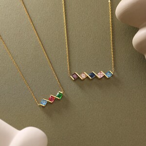 May include: Two gold necklaces with colorful square gemstones. The necklaces are on a green background. The top necklace has five gemstones in a diagonal line: pink, blue, dark blue, pink, and light blue. The bottom necklace has four gemstones in a diagonal line: light blue, red, light blue, and green.
