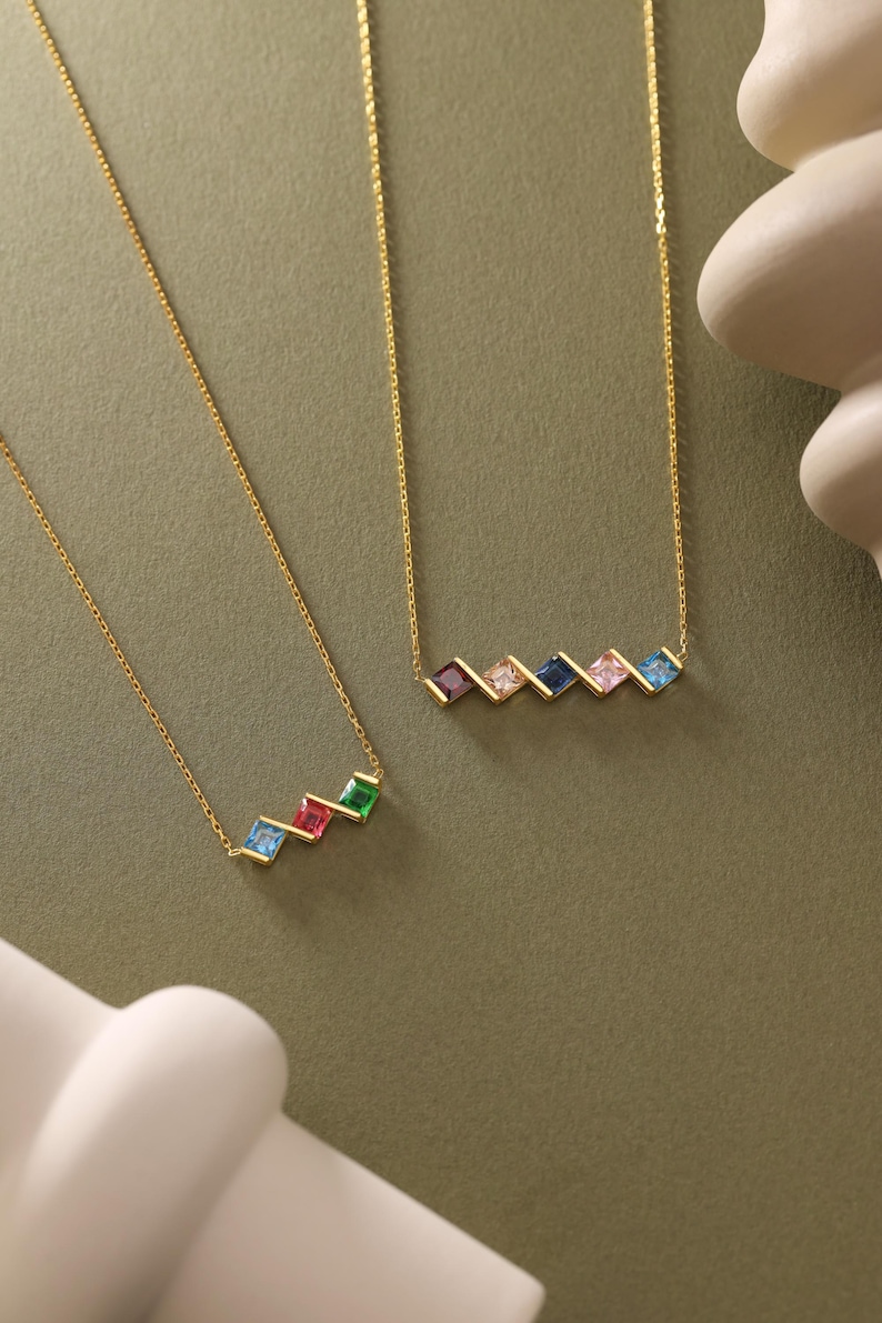 May include: Two gold necklaces with colorful square gemstones. The necklaces are on a green background. The top necklace has five gemstones in a diagonal line: pink, blue, dark blue, pink, and light blue. The bottom necklace has four gemstones in a diagonal line: light blue, red, light blue, and green.