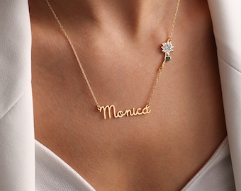 May include: A gold necklace with a delicate chain and a pendant featuring the name "Monica" in cursive script. A small flower charm with green leaves hangs on the side of the chain.
