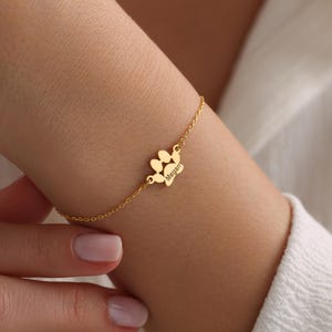 Puede incluir: Pulsera de cadena dorada con un dije de huella de pata grabado con el nombre "Megan".