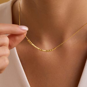 Peut inclure: Un délicat collier en or composé d'une série de petites perles et de barres cylindriques. Le collier est porté autour du cou et est tenu par une main. Un bijou simple et élégant.