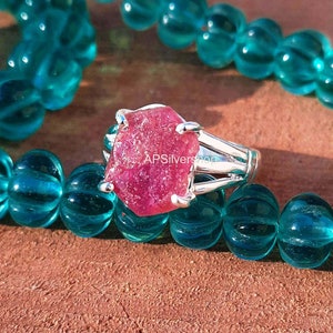Könnte beinhalten: Ein Silberring mit einem großen, rohen, rosafarbenen Rubin-Edelstein. Der Ring ist vor einem Hintergrund aus türkisfarbenen Perlen platziert.