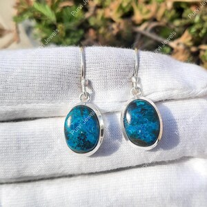 May include: A pair of silver drop earrings featuring oval, blue-green stones. The stones have a marbled effect, set in a silver bezel. The earrings are suspended from a simple silver hook.