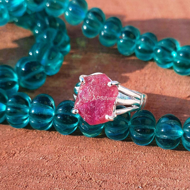 K&ouml;nnte beinhalten: Ein Silberring mit einem gro&szlig;en, rohen, rosafarbenen Rubin-Edelstein. Der Ring ist vor einem Hintergrund aus t&uuml;rkisfarbenen Perlen platziert.