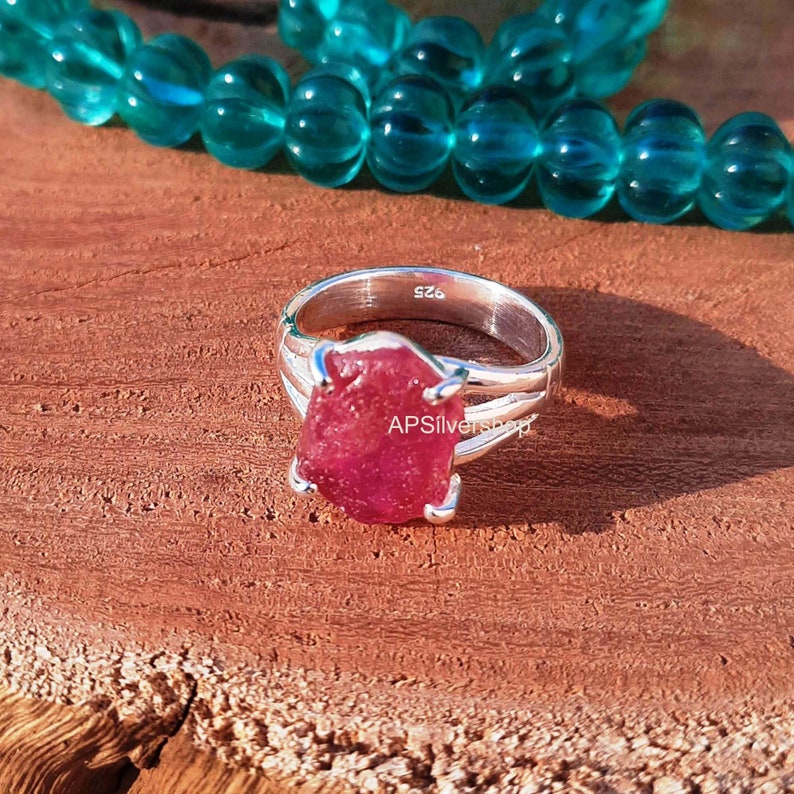 K&ouml;nnte beinhalten: Ein silberner Ring mit einem gro&szlig;en, rohen, rosafarbenen Rubin-Edelstein, der in der Mitte eingesetzt ist. Der Ring hat ein schlichtes, elegantes Design mit zwei B&auml;ndern, die um den Stein gewickelt sind. Der Ring ist Gr&ouml;&szlig;e 6.